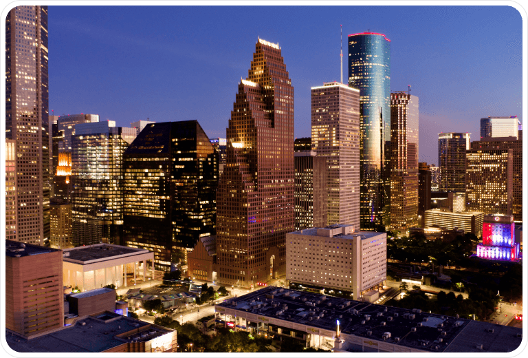 City skyline at dusk with illuminated skyscrapers and a clear sky, showcasing modern architecture and vibrant lighting.